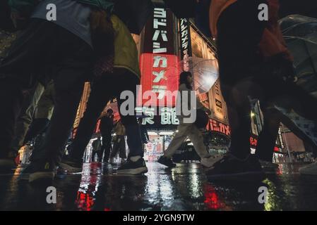 Leute mit Sonnenschirmen laufen abends bei Regen am Yodobashi Kamera Shop in Shinjuku vorbei. Die Neonlichter werden auf der nassen Straße reflektiert Stockfoto