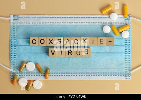 Coxsackievirus. Die Worte sind mit Holzwürfeln auf einer chirurgischen Gesichtsmaske angeordnet. Es liegen verschiedene Pillen herum. Stockfoto