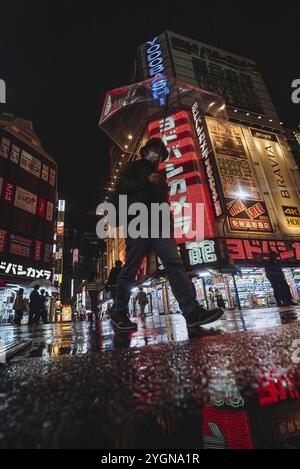 Ein Mann mit einem Schirm geht abends bei Regen am Yodobashi Kamera Shop in Shinjuku vorbei. Die Neonlichter werden auf der nassen Straße reflektiert Stockfoto