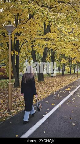 Eine ästhetische junge Frau mit schwarzem Mantel spaziert durch die bunten Baumallee im Herbstpark. Ruhige Herbstszene Stockfoto