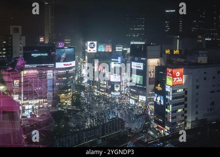 Die Menschen laufen abends im Regen über die berühmte Shibuya Scramble Crossing, beleuchtet durch Werbung und große Bildschirme Stockfoto