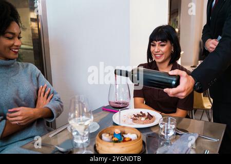Verschiedene Freundinnen genießen Weinverkostungen in einem gehobenen Restaurant mit Sommelier, der Rotwein serviert Stockfoto