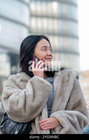 Eine schöne 20-25-jährige Frau in einem weichen orientalischen Mantel spricht an einem hellen Tag mit ihrem Smartphone und lächelt vor dem Hintergrund einer modernen Stadt. Stockfoto