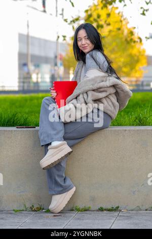 Eine junge Frau von 20-25 Jahren orientalischem Aussehen sitzt auf einer niedrigen Wand mit einem Ordner in der Hand und genießt einen sonnigen Tag in einem Stadtpark. Stockfoto