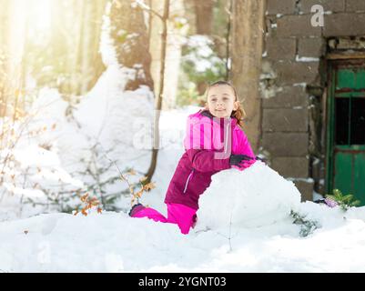 Glückliches Kind, das während der Winterferien in einem verschneiten Wald in der Nähe eines alten verlassenen Hauses einen Schneemann macht Stockfoto