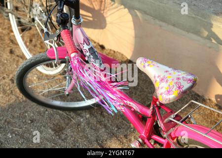 Rosafarbenes Fahrrad mit bunten Quasten und geblümtem Sattel, das neben einer Mauer geparkt ist und an einem sonnigen Tag einen Schatten wirft Stockfoto