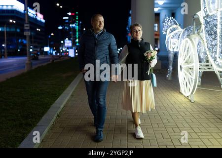 Ein Paar spaziert bei Nacht Hand in Hand durch die Stadt, vorbei an einer beleuchteten Kutsche Stockfoto