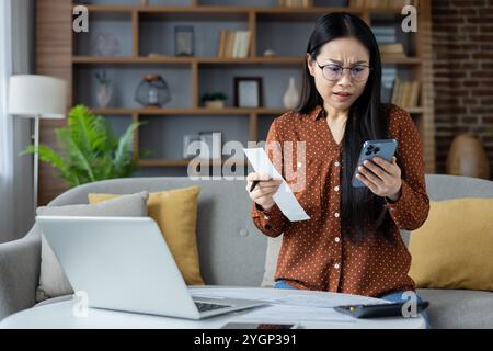 Asiatische Frau, die sich gestresst fühlt, während sie Rechnungen zu Hause mit Smartphone und Laptop verwaltet. Sie hält die Rechnung mit besorgter Ausdruckskraft und steckt in Online-Banking-Aufgaben. Finanz- und Technologiekonzept. Stockfoto