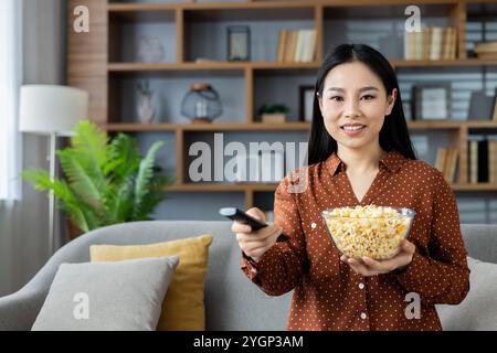 Asiatische Frau, die bequem auf dem Sofa sitzt, Popcorn und Fernbedienung hält. Entspannte Atmosphäre zu Hause mit gemütlichen Kissen, die auf Freizeit schließen lassen. Bereit für den Filmabend, genießt sie drinnen Ruhe. Stockfoto