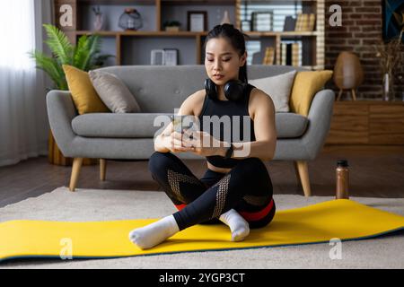 Asiatische Frau in Trainingskleidung mit Smartphone und Kopfhörern während des Heimtrainings auf Yogamatte. Engagierte sich für Fitnessaktivitäten, die zeitgenössischen Lebensstil und Achtsamkeitspraktiken widerspiegeln. Stockfoto