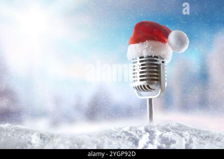 Vintage-Mikrofon mit Weihnachtsmann-Hut und Metallständer auf einem Schneehaufen mit einer verschneiten Landschaft im Hintergrund. Vorderansicht. Stockfoto