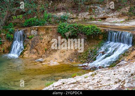 Ein kleiner Wasserfall fließt sanft über felsiges Gelände, umgeben von üppigem Grün und einem ruhigen Pool darunter. Stockfoto