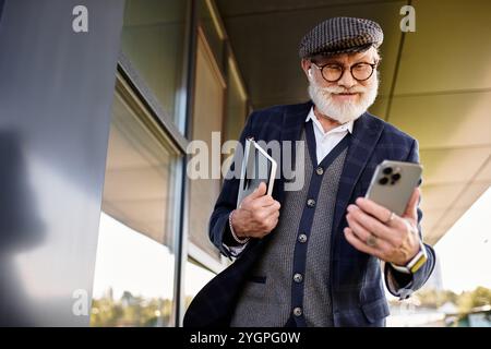 Ein stilvoller Seniorenmann mit Bart überprüft sein Smartphone, während er im Herbst in der Nähe eines modernen Gebäudes steht. Stockfoto