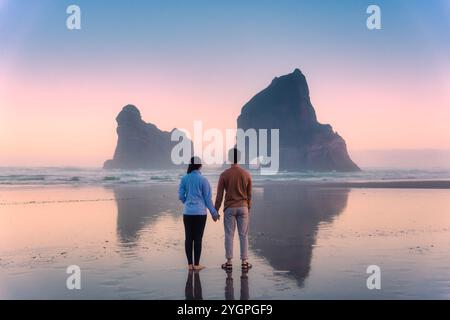 Ein junges Paar, das mit Händen steht und den Wharariki Beach und die Archway Inseln auf der Tasmanischen See im Westen von Cape Farewell, Neuseeland, genießt Stockfoto