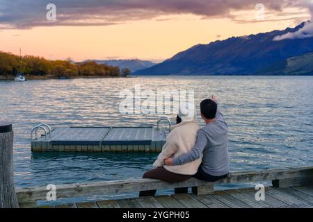 Rückansicht eines asiatischen Paares, das im Herbst auf dem Lake Wakatipu in Neuseeland sitzt und den Sonnenuntergang und den Berg genießt Stockfoto