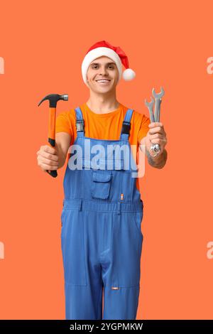 Männlicher Klempner im Weihnachtsmann-Hut mit Hammer und Schraubenschlüsseln auf orangem Hintergrund Stockfoto
