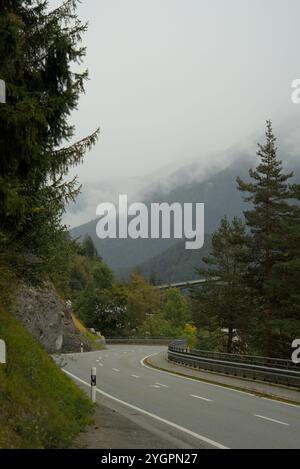 Gewundene Bergstraße durch die Schweizer Alpen an einem nebeligen Tag mit dichtem Grün und bewölktem Himmel Stockfoto