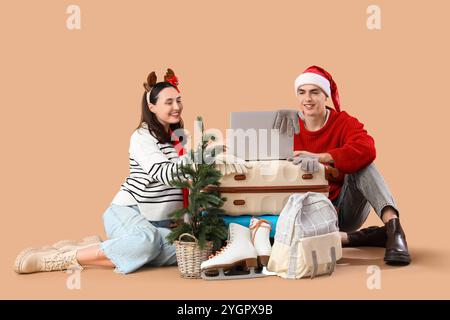 Nettes junges Paar mit Laptop, Koffern und kleinem Weihnachtsbaum auf beigefarbenem Hintergrund. Winterurlaubskonzept Stockfoto