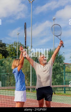 Das Tennis-Paar der vertikalen Senioren hebt seine Schläger hoch und jubelt fröhlich an, um ihren Sieg in einem Spiel zu feiern. Ihre enthusiastischen Ausdrücke spiegeln sich wider Stockfoto