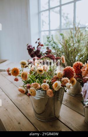 Helle und farbenfrohe Blumen in Metalleimern zieren einen Holztisch am Fenster und schaffen eine rustikale und ruhige Atmosphäre Stockfoto