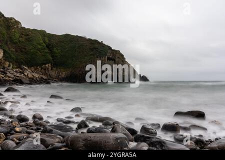 Lange Exposition von nebeligen Meereswellen, die über Felsen am Strand mit Klippen und natürlichem Bogen im Hintergrund Stockfoto