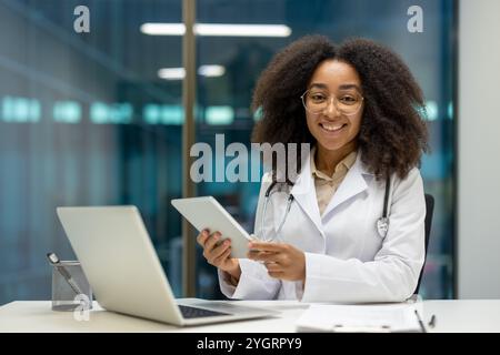Lächelnde afroamerikanische Ärztin mit Tablet im Büro. Das Tragen von weißem Mantel und Stethoskop, die Technologie im Gesundheitswesen darstellen. Fördert moderne Kommunikation und Patientenbindung. Stockfoto