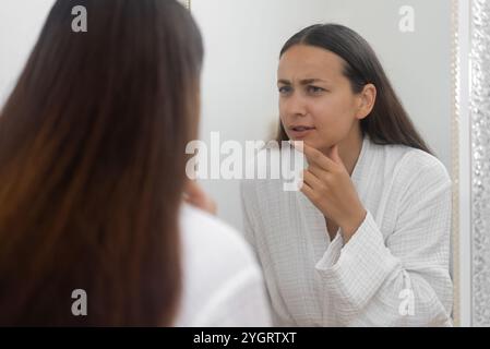 Im Badezimmer schaut eine besorgte Frau mit Traurigkeit auf ihr Spiegelbild, beunruhigt von Hautproblemen Stockfoto