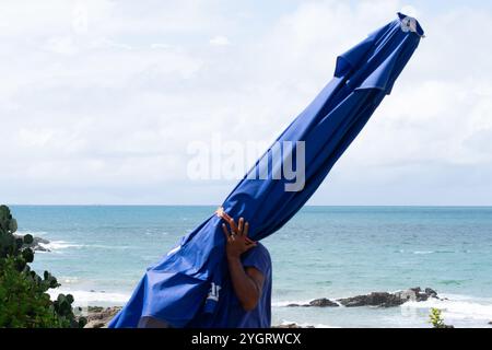 Salvador, Bahia, Brasilien - 11. Oktober 2019: Ein Strandarbeiter wird bei starker Sonneneinstrahlung am Strand Barra in der Stadt Salva mit einem blauen Sonnenschirm gesehen Stockfoto