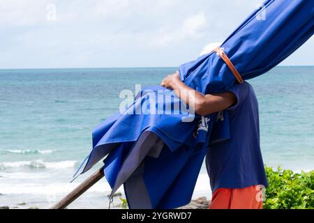 Salvador, Bahia, Brasilien - 11. Oktober 2019: Ein Strandarbeiter wird bei starker Sonneneinstrahlung am Strand Barra in der Stadt Salva mit einem blauen Sonnenschirm gesehen Stockfoto