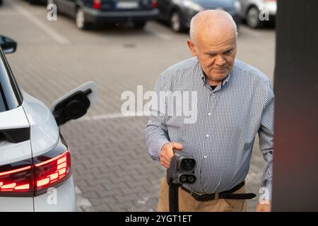 Ein älterer Mann hält einen Ladestecker für Elektrofahrzeuge und bereitet sich darauf vor, ihn auf einem Parkplatz an sein Auto anzuschließen, um eine nachhaltige Energienutzung zu fördern. Stockfoto