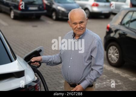 Ein älterer Mann lächelt in die Kamera, während er ein Ladekabel hält, das mit seinem Elektroauto verbunden ist, was einen Schritt hin zu umweltfreundlichen Verkehrsmitteln darstellt Stockfoto