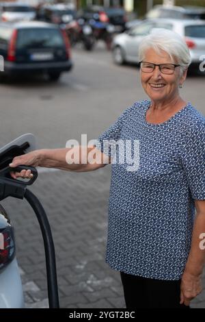 Eine ältere Frau lächelt freundlich in die Kamera, während sie ein Ladekabel hält, das mit ihrem Elektrofahrzeug verbunden ist, was einen nachhaltigen Energieverbrauch symbolisiert. Stockfoto