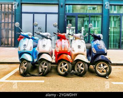 Eine Reihe von vier Motorrollern, die jeweils in hellen Farben wie Blau, Rot, Silber und Hellblau lackiert sind, parkt dicht beieinander auf einer Stadtstraße. Sie A Stockfoto