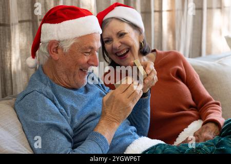 Weihnachten, Seniorenpaar mit Weihnachtsmützen, die Geschenke austauschen und auf einem gemütlichen Sofa lachen, zu Hause Stockfoto