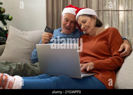 Weihnachten, Seniorenpaar, das online mit Laptop einkaufen will, weihnachtsmütze trägt, zu Hause Stockfoto