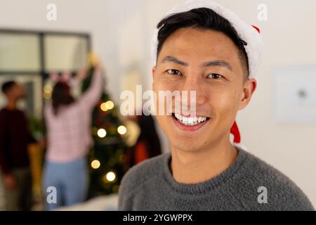 Weihnachtszeit, lächelnder Mann mit weihnachtsmann-Hut mit multirassischer Familie, die Baum dekoriert, zu Hause Stockfoto