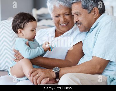 Spielzeug, Lächeln und Großeltern mit Baby auf dem Sofa zum Binden, Spielen und Stützen. Liebe, Glück und Generationen mit älteren Männern und Frauen in der Hand Stockfoto