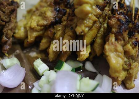 Nahaufnahme gegrillter Hähnchensatay-Spieße mit reichhaltiger Erdnusssauce, frischer Gurke und Zwiebelscheiben. Stockfoto
