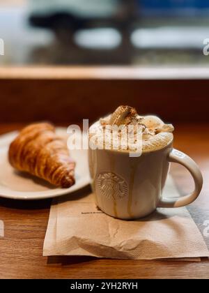 „Eine gemütliche Morgenszene mit einem schaumigen Cappuccino mit Schlagsahne und Zimt sowie einem knusprigen Croissant auf einem Holztisch.“ Stockfoto