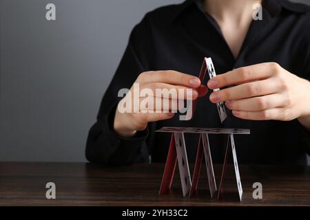 Frau baut Haus mit Spielkarten an Holztisch vor grauem Hintergrund, Nahaufnahme. Leerzeichen für Text Stockfoto