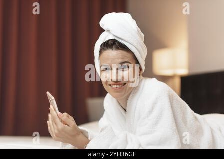 Junge Frau im Bademantel im Zimmer über Handy liegen, entspannt nach einem Bad Stockfoto