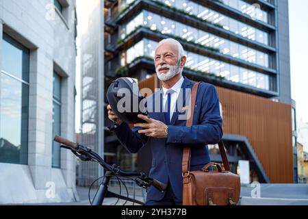 Senior männlicher Pendler, der Helm aufsetzt, während er auf der Straße vor dem modernen Gebäude steht Stockfoto