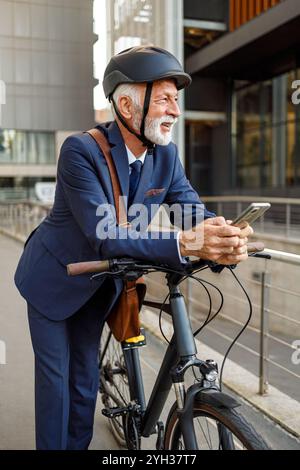 Nachdenklicher männlicher Profi im Helm, der das Telefon benutzt und wegblickt, während er sich in der Stadt auf das Fahrrad lehnt Stockfoto