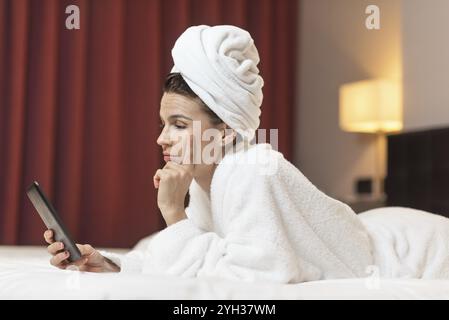 Junge Frau im Bademantel im Zimmer über Handy liegen, entspannt nach einem Bad Stockfoto