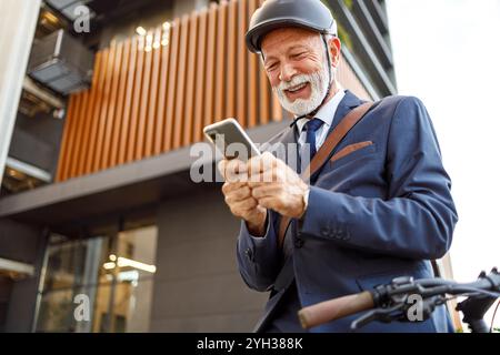 Niedriger Blickwinkel auf einen fröhlichen älteren Mann im Helm, der während der Fahrt in der Stadt über das Mobiltelefon Nachrichten sendet Stockfoto
