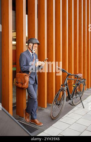 In voller Länge ein leitender Geschäftsmann, der über Handy SMS schreibt, während er neben dem Fahrrad an einer Holzwand steht Stockfoto