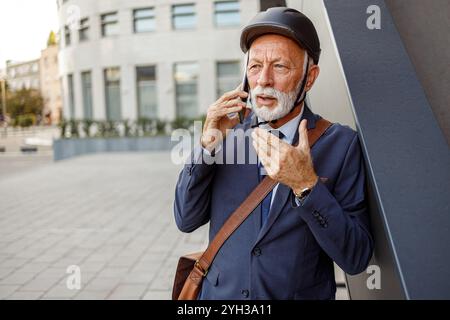 Porträt eines seriösen Geschäftsmannes mit Helm, der über das Smartphone hört, während er in der Stadt an der Wand steht Stockfoto