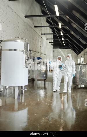 Vertikale Aufnahme einer professionellen afroamerikanischen Supervisorin in Maske und Overall, die die Werkstattausrüstung inspiziert, die auf den Tank zeigt, während sie darüber diskutieren Stockfoto