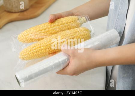 Frau, die Plastikfutterverpackung über Maiskolben in der Küche legt, Nahaufnahme Stockfoto