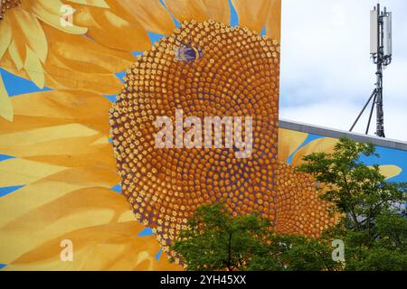 Ein großes Wandbild einer Sonnenblume bedeckt ein Gebäude, dessen Zentrum ein Fibonacci-Muster zeigt. Neben dem vib steht ein Sendeturm Stockfoto